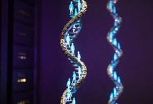 An artistic, vertical depiction of a DNA double helix designed like an ancient, spiraling staircase. Small, glowing cyan figures of people are standing and walking along the rungs of the DNA strand. The helix itself has an intricate, carved stone or metal texture, set against a dark, moody violet background with soft bokeh lights in the distance. Artistic illustration for Genome India Project